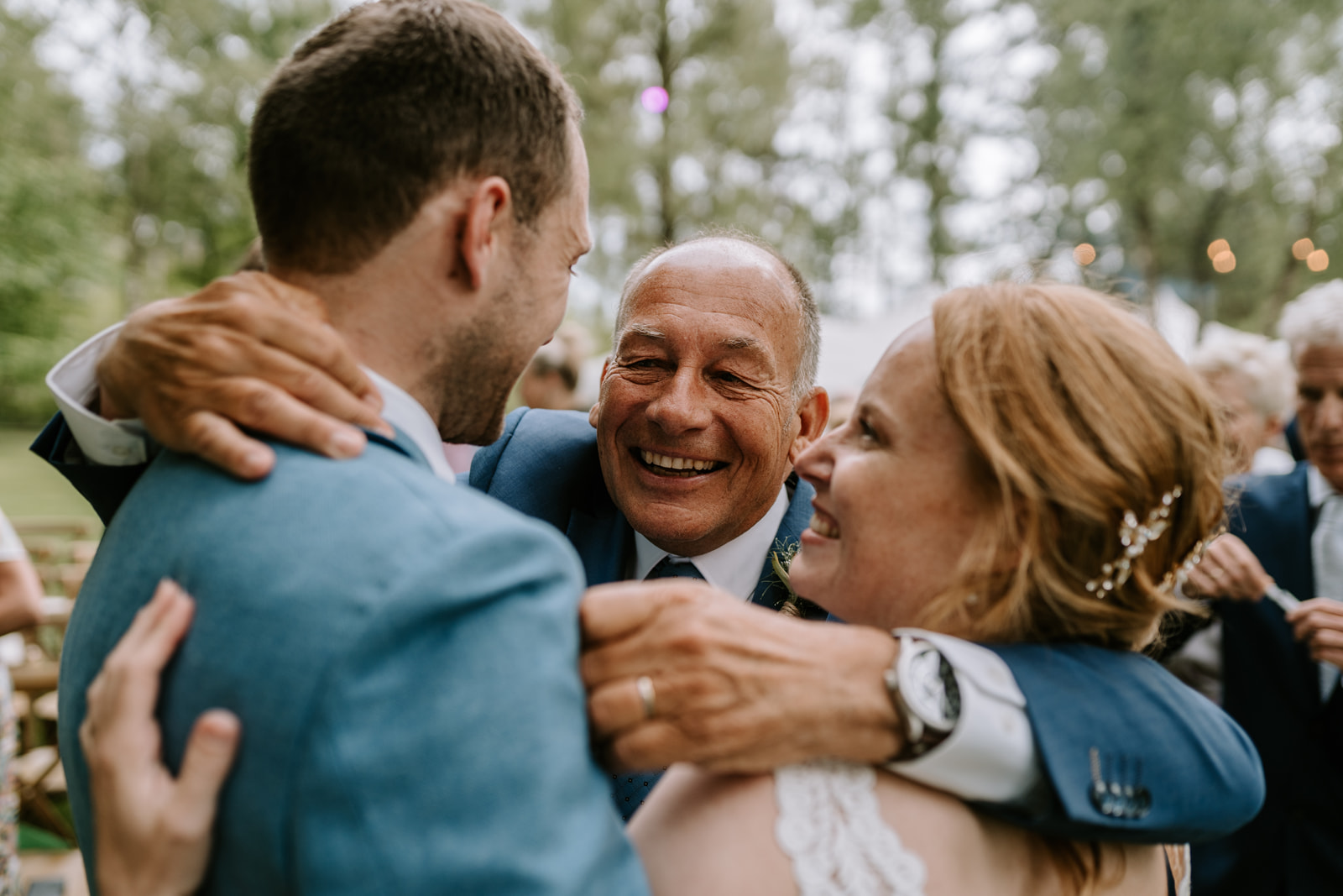De vader van de bruidegom geeft het bruidspaar een innige knuffel met het bos en gasten op de achtergrond.